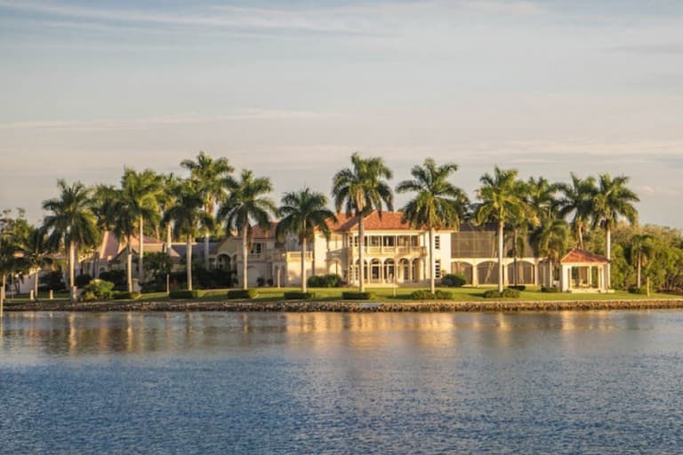 Looking onto a large mansion with palm trees outside surrounded by water